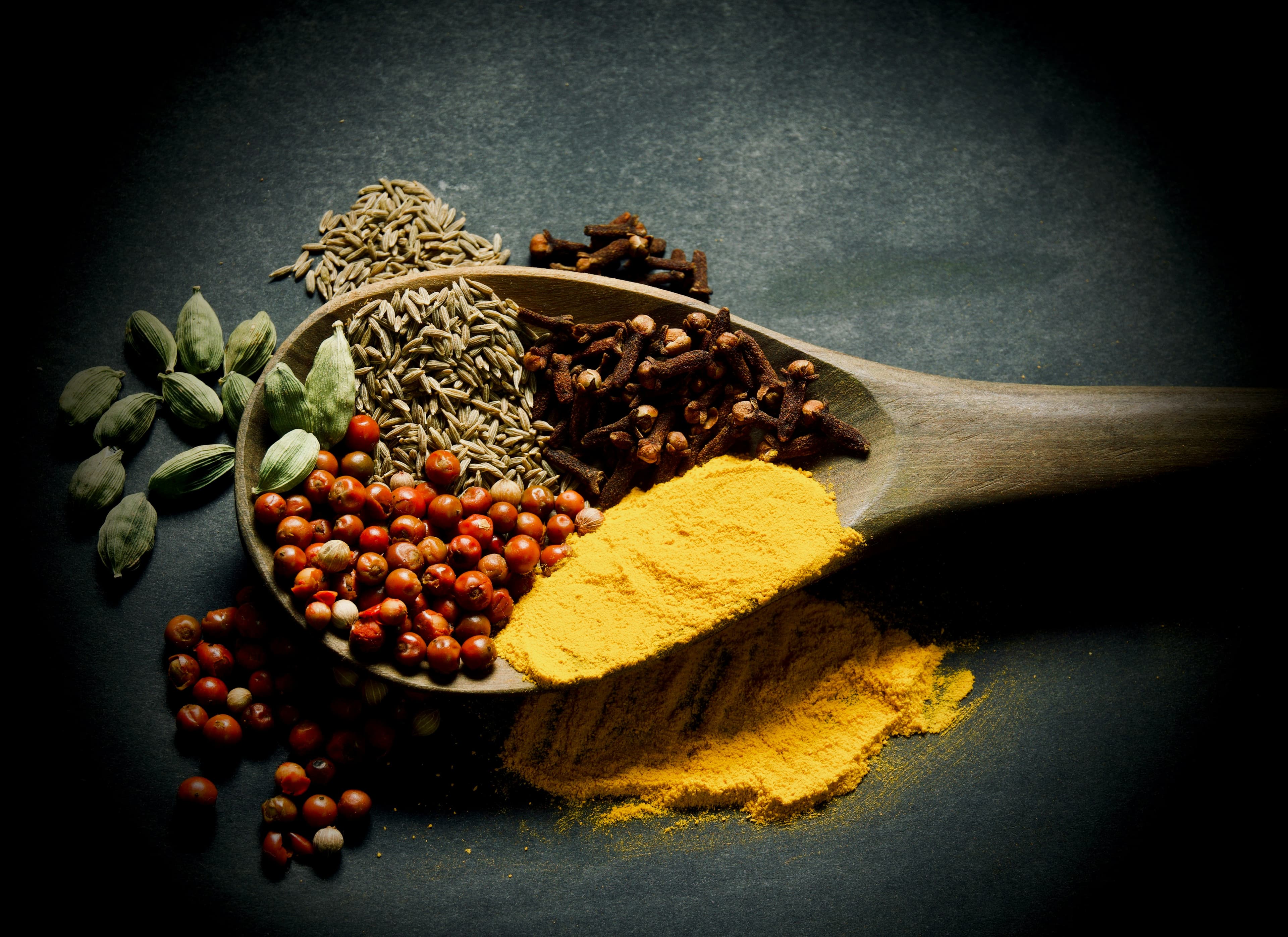 A wooden spoon filled with whole and ground spices including turmeric, cardamom, and cumin