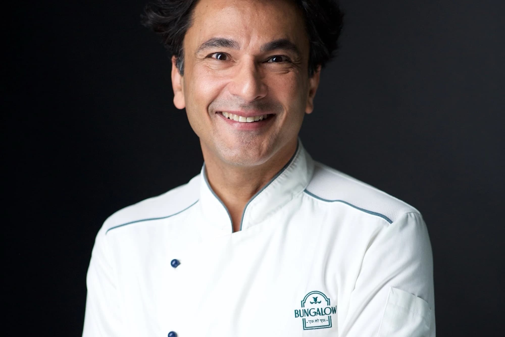 Chef Vikas Khanna in a white Bungalow chef coat, smiling against a black studio background